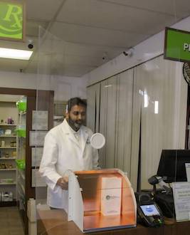 Pharmacist behind protective shield. He is using a copper-lined port to deliver medications to customers.